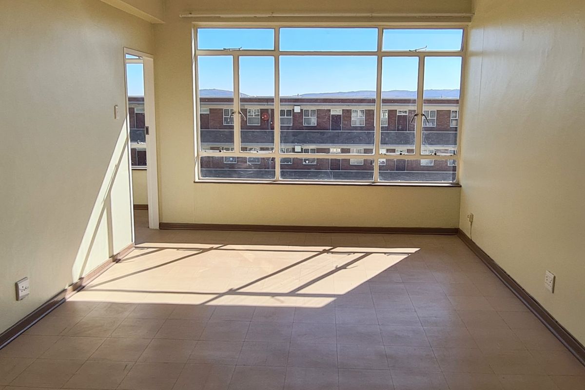 A bright, empty living area featuring large windows, tiled flooring, and neutral wall finishes, offering a blank canvas for potential renovation or interior design.