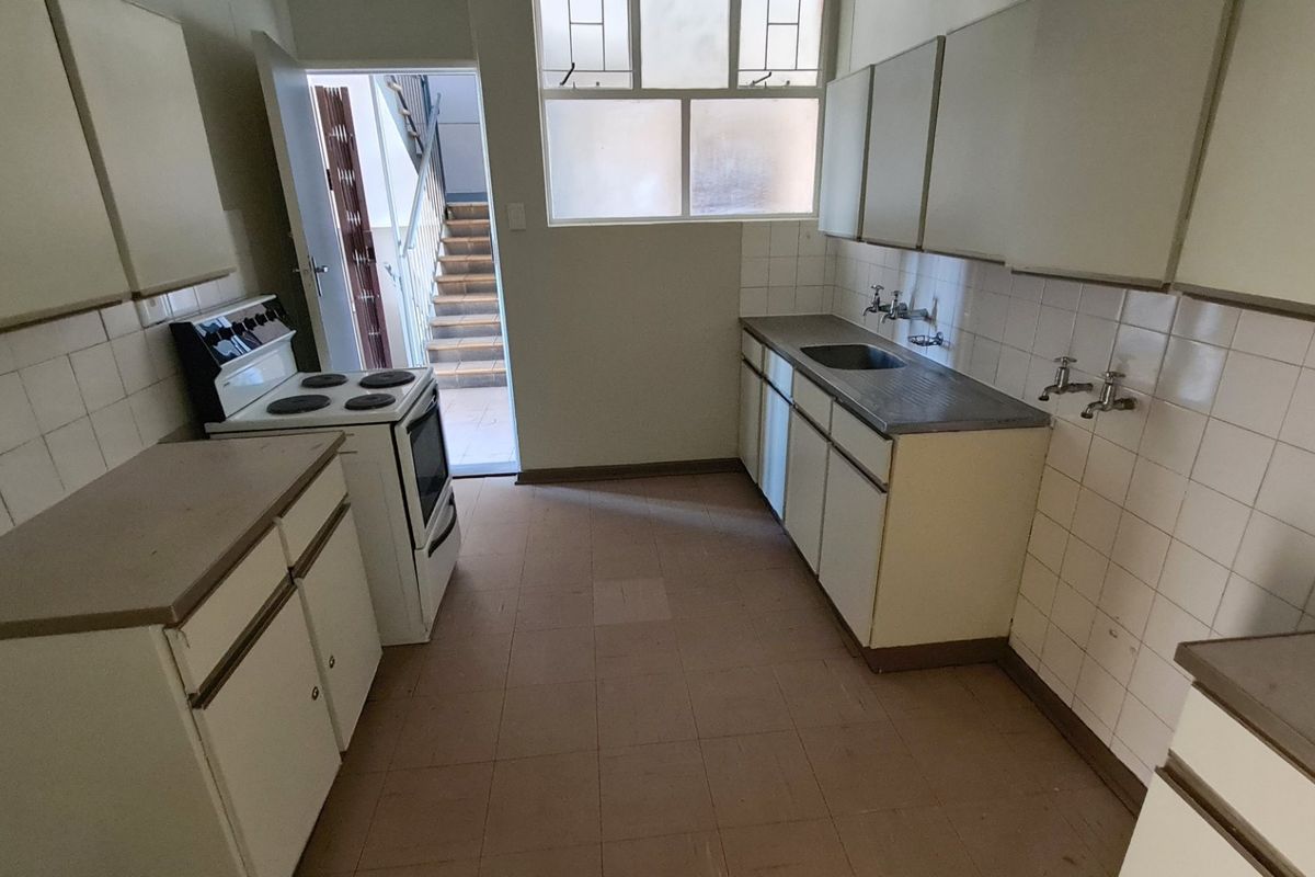 A functional, dated kitchen featuring built-in cabinetry, a stainless steel sink, tiled splashbacks, and a freestanding stove, with direct access to an external stairwell.