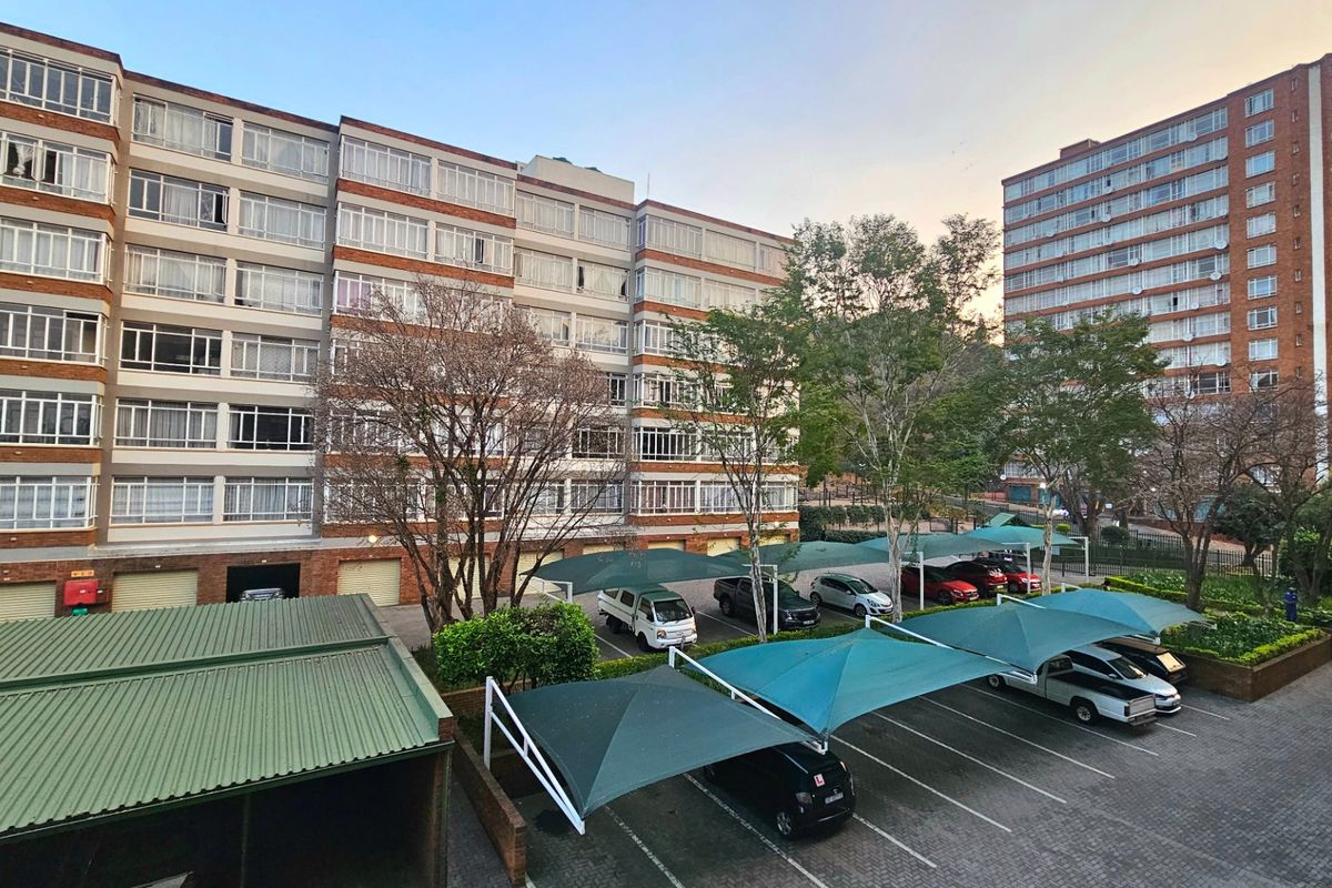 Exterior view of a multi-story commercial or residential building featuring a paved parking area with shade cloth carports and established landscaping.
