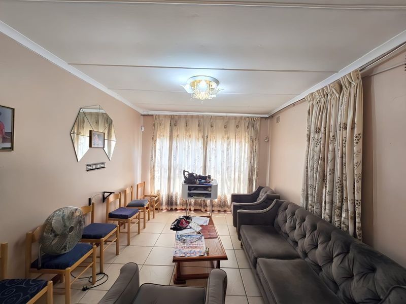 A living room featuring tiled flooring and a central ceiling light fixture, with large windows covered by curtains.