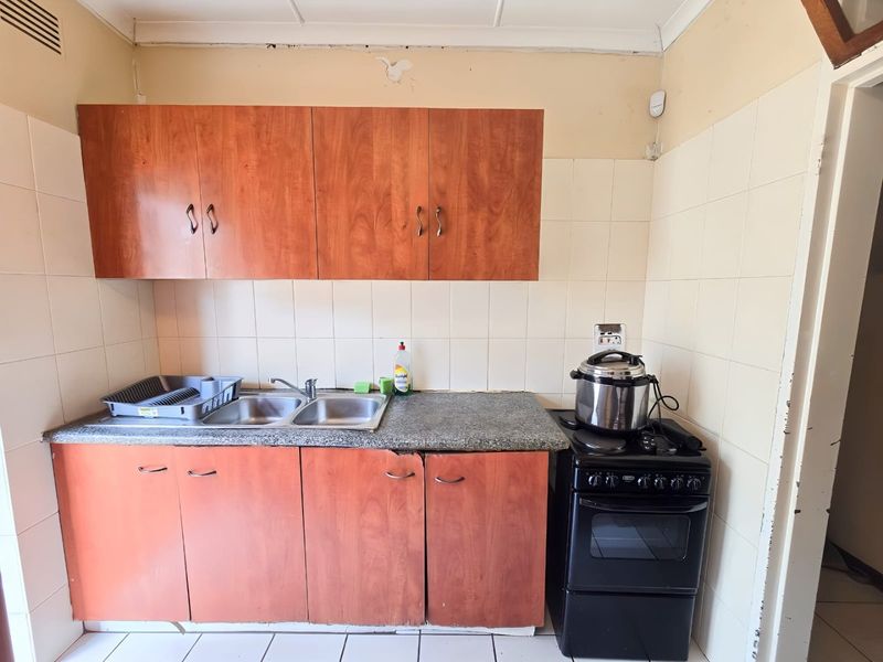 A compact kitchen featuring wooden cabinetry, a double stainless steel sink, and tiled surfaces, currently showing signs of age and wear.