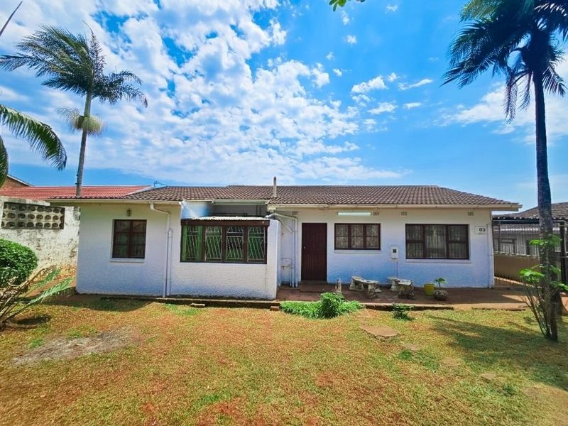 Rear view of a single-story house featuring a tiled roof, security bars on windows, and a concrete patio area overlooking the garden.