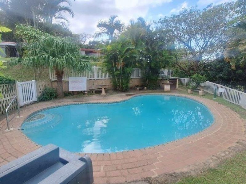 A communal swimming pool area featuring a kidney-shaped pool, brick coping, and surrounding landscaped garden with white picket fencing.
