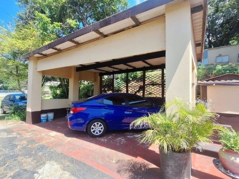 A covered carport structure providing sheltered parking, featuring concrete pillars and a metal roof, situated on a paved driveway area.