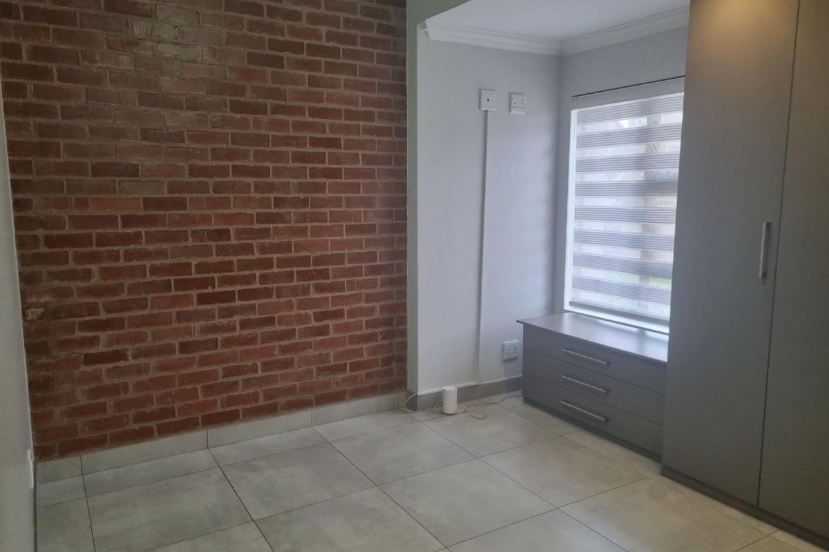 A bedroom featuring a distinctive exposed brick feature wall, neutral floor tiling, built-in grey cabinetry, and a window with modern zebra blinds.