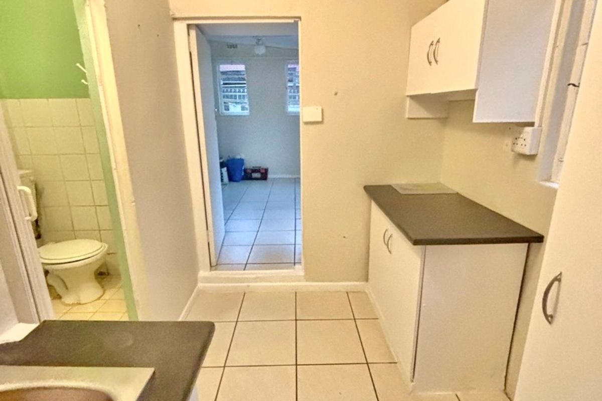 A compact kitchen area featuring built-in cabinetry, laminate countertops, and tiled flooring, with an adjacent bathroom and doorway leading to another room.