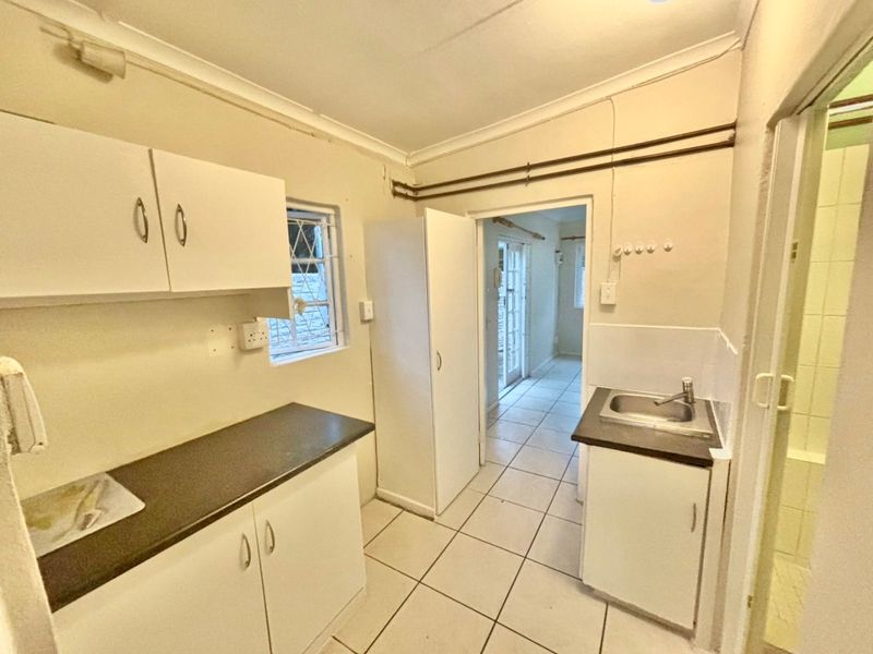 A compact, empty kitchen featuring white built-in cabinetry, dark laminate countertops, a stainless steel sink, and tiled flooring with visible surface-mounted electrical conduits.