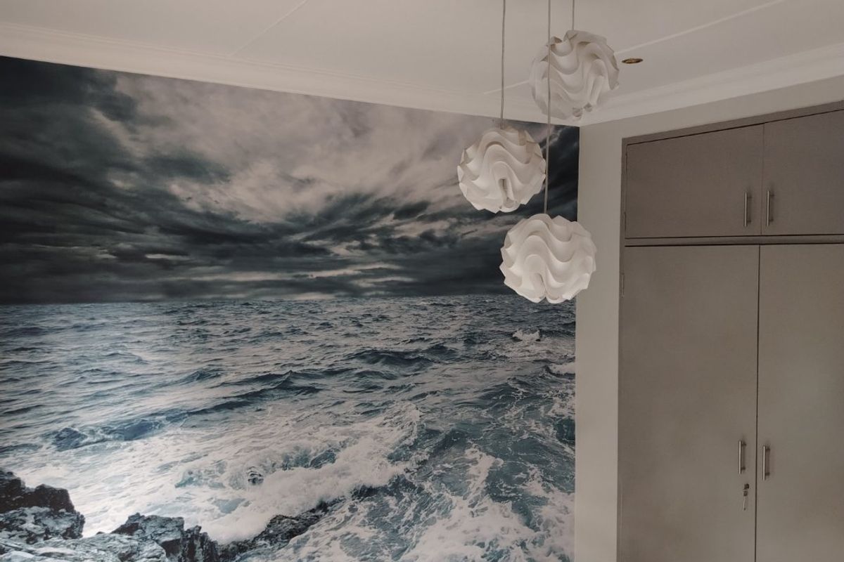 A bedroom featuring a striking ocean-themed feature wall, built-in grey cabinetry, light wood-look flooring, and modern pendant lighting.