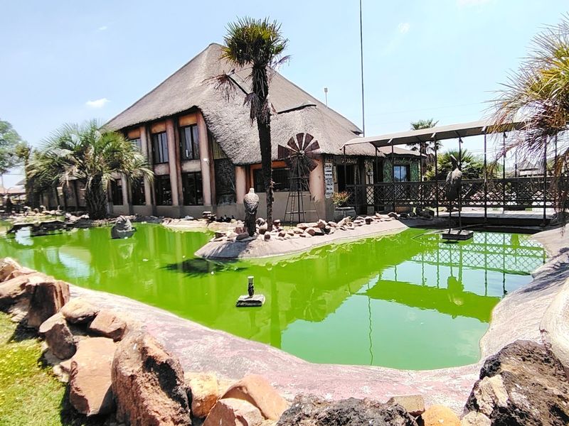 Exterior view of a commercial property featuring a large ornamental pond, stone landscaping, a pedestrian bridge, and a distinctive thatched roof structure.
