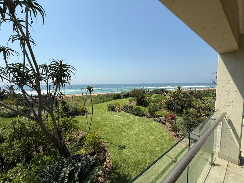 A scenic view from a balcony overlooking a landscaped garden with lush greenery and a direct view of the ocean and beach.
