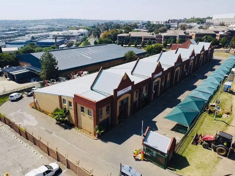 Aerial view of an industrial warehouse complex featuring multiple units, pitched metal roofing, brick construction, a security gatehouse, and shaded parking facilities.