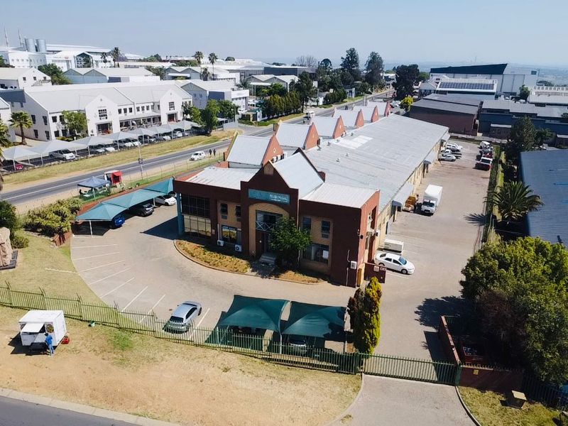 Aerial view of an industrial property featuring a main building with metal roofing, extensive asphalt parking, and perimeter security fencing.