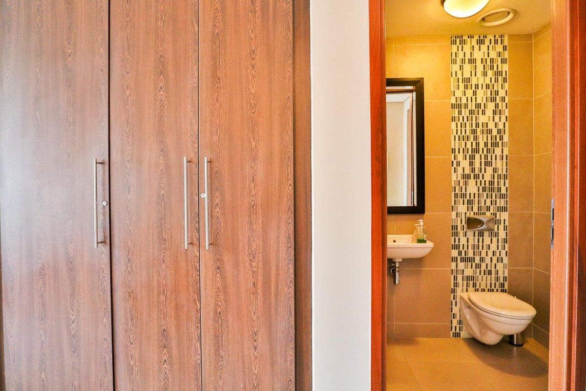 A clean bathroom featuring a wall-mounted toilet, basin, and a decorative mosaic tile accent wall, adjacent to built-in wooden storage cupboards.