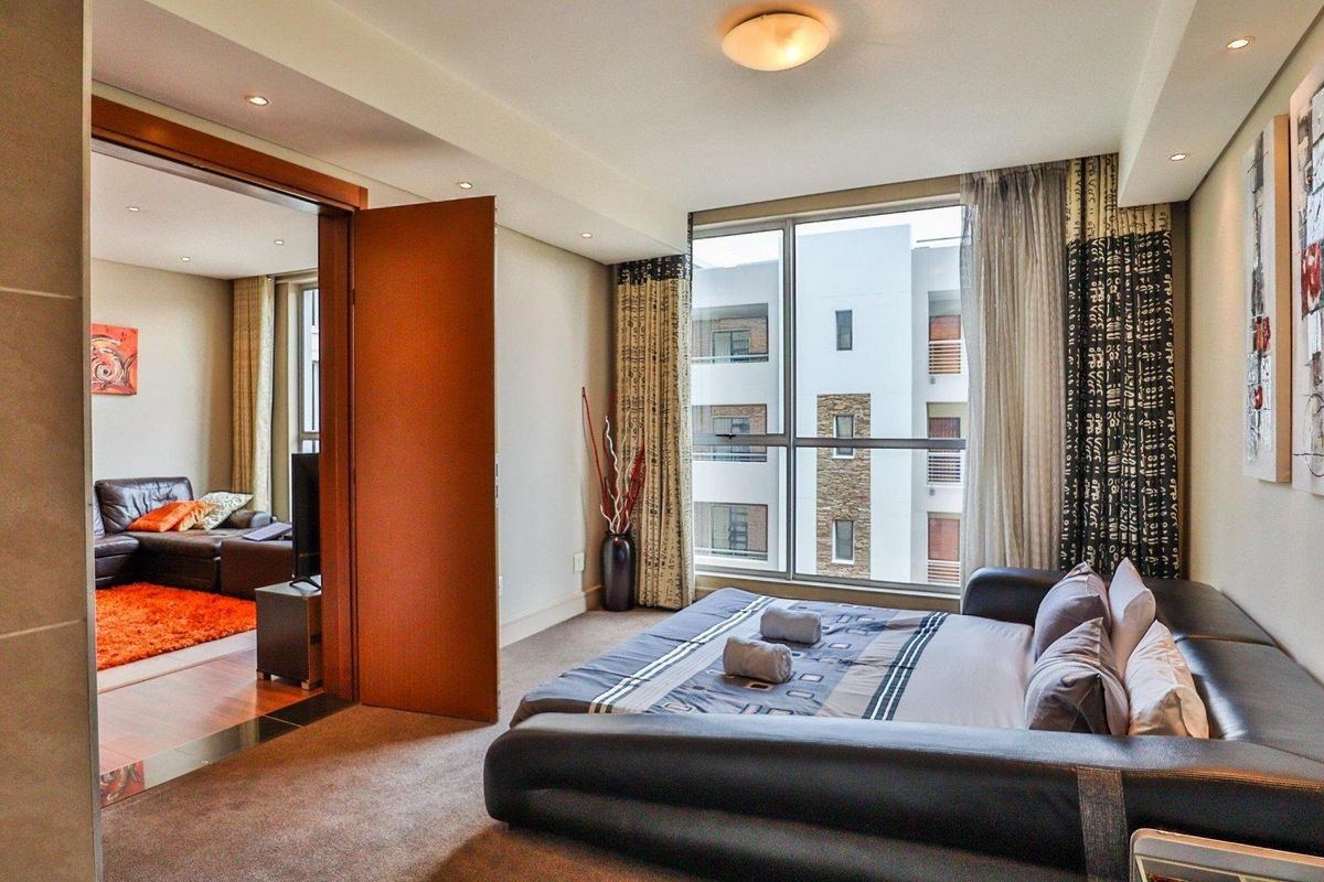 A well-lit bedroom featuring large floor-to-ceiling windows, neutral carpeting, and recessed lighting, with an open doorway leading into an adjacent living area.