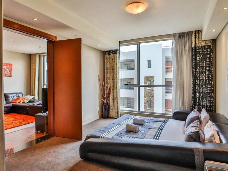 A well-lit bedroom featuring large floor-to-ceiling windows, neutral carpeting, and recessed lighting, with an open doorway leading into an adjacent living area.