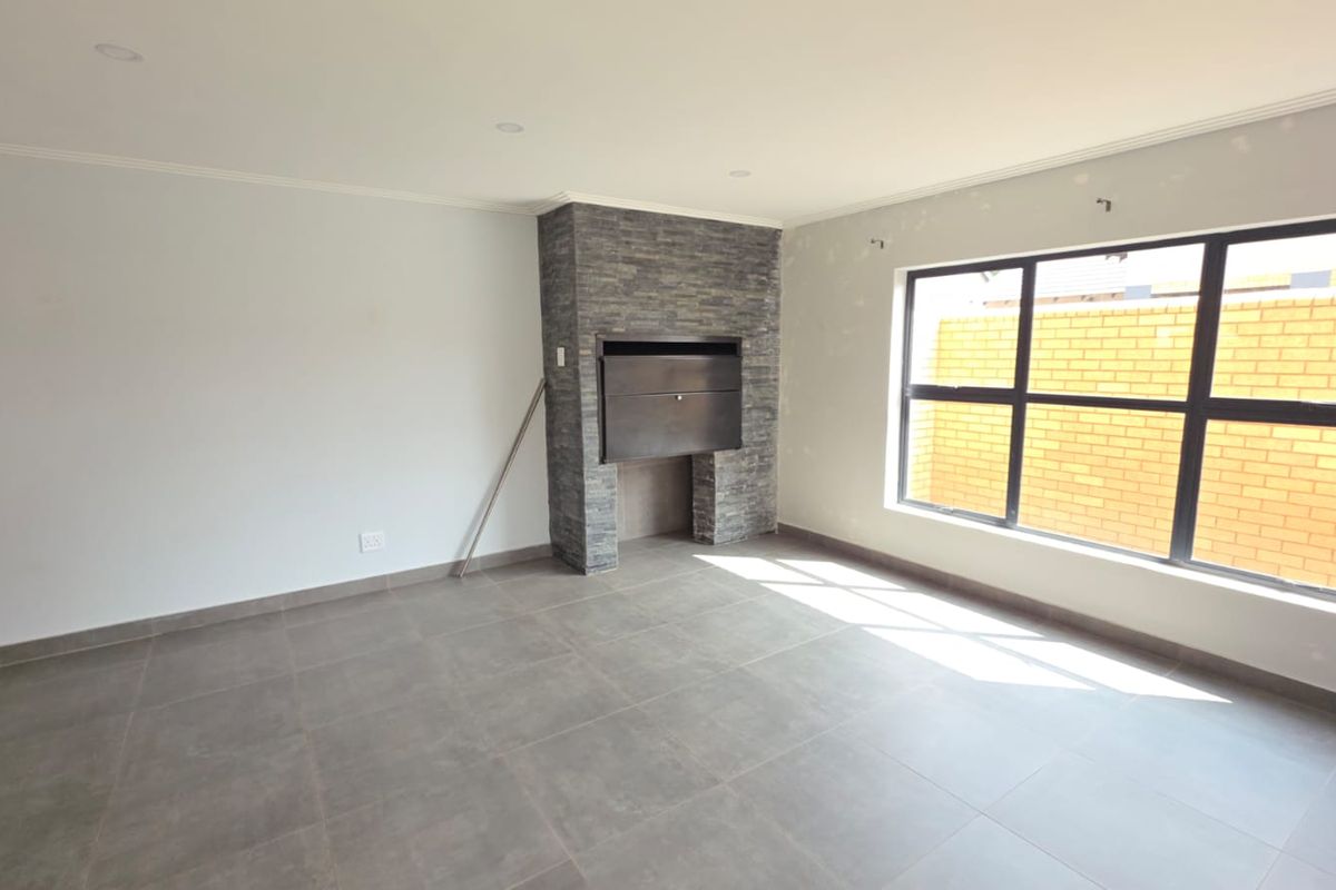 A bright, empty living room featuring large windows, tiled flooring, and a built-in stone-clad fireplace, currently awaiting final finishing touches.