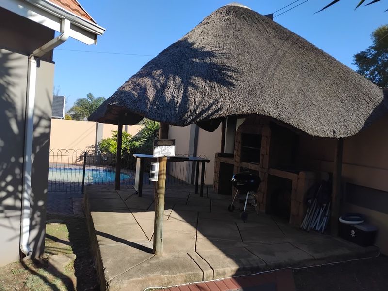 Outdoor thatched lapa area featuring a built-in braai and concrete patio, overlooking the swimming pool.