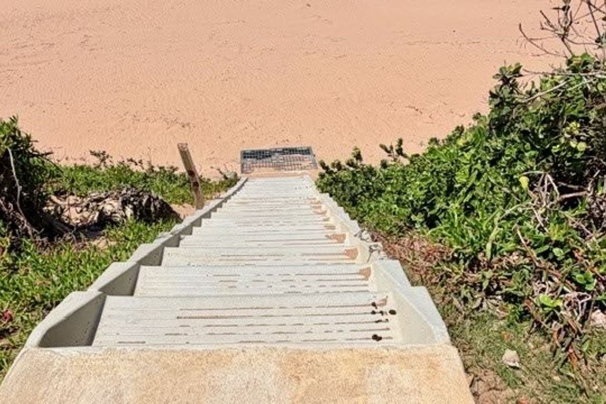A set of concrete stairs providing direct access to the beach, surrounded by natural coastal vegetation.