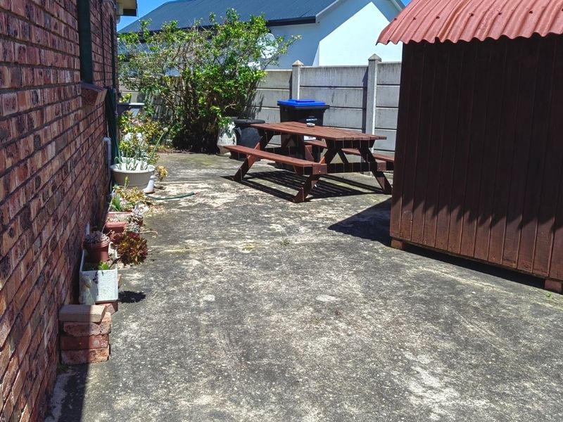 A rear exterior area featuring a brick house wall, concrete paving, and a small storage shed with a corrugated metal roof.
