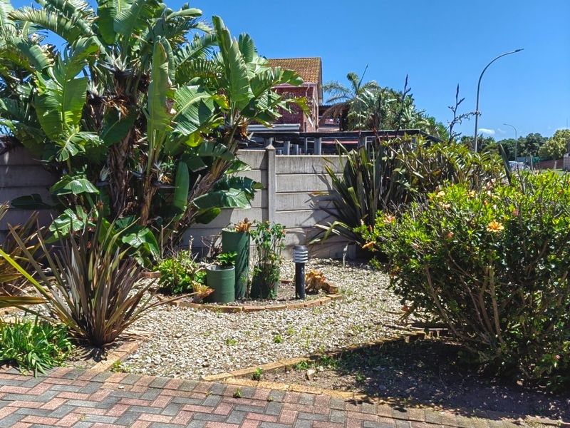 A landscaped front garden area featuring established tropical plants, a gravel bed, and a paved driveway with a precast concrete boundary wall.