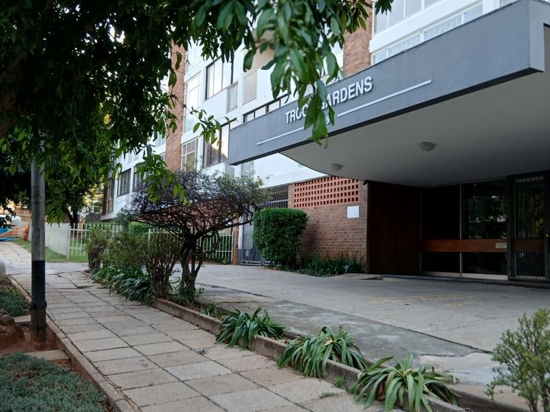 Front exterior view of the apartment building entrance, featuring a covered canopy, brick facade, and paved walkway access.