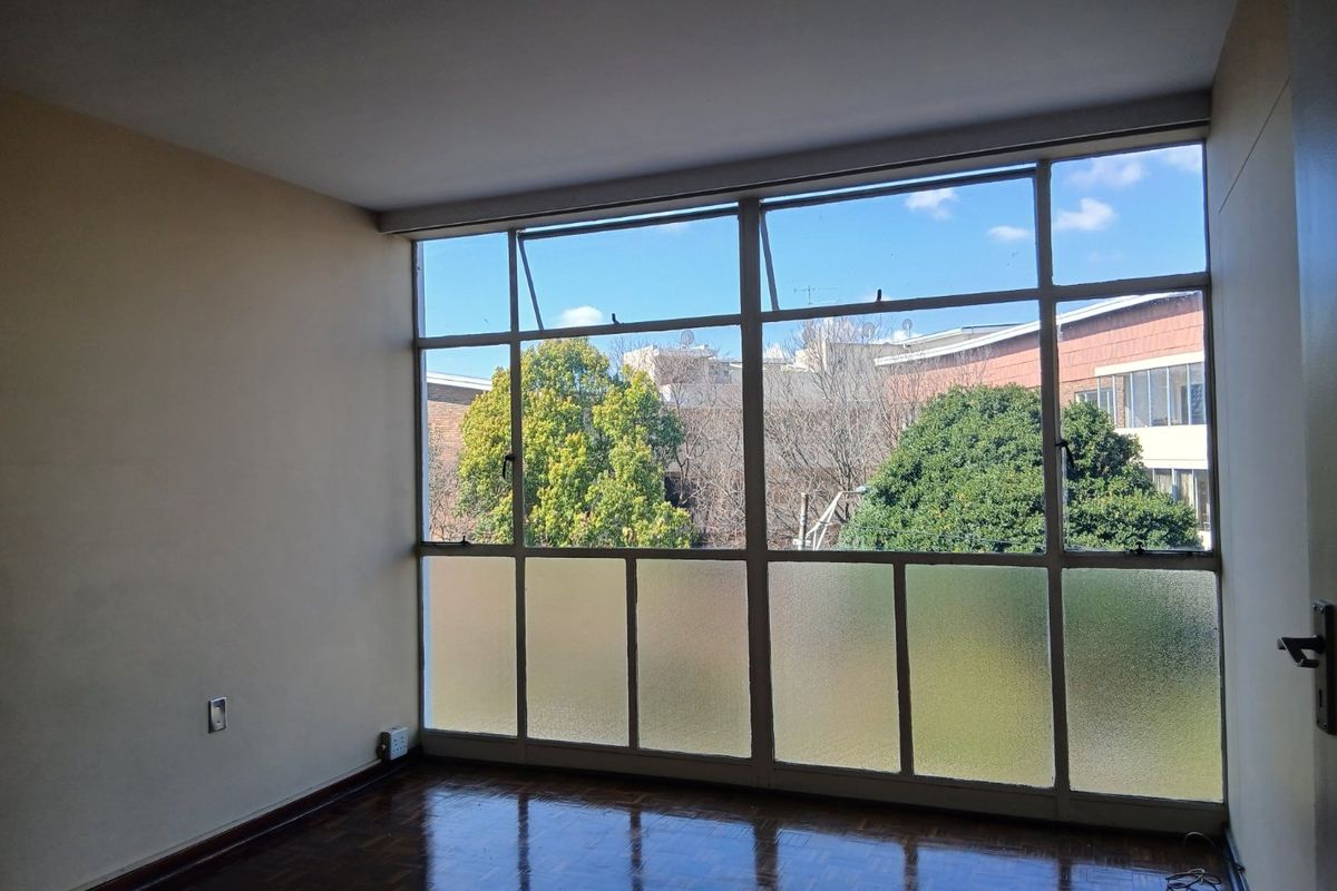 An empty bedroom featuring original parquet flooring and large metal-framed windows with frosted glass lower panels, offering natural light and an urban outlook.