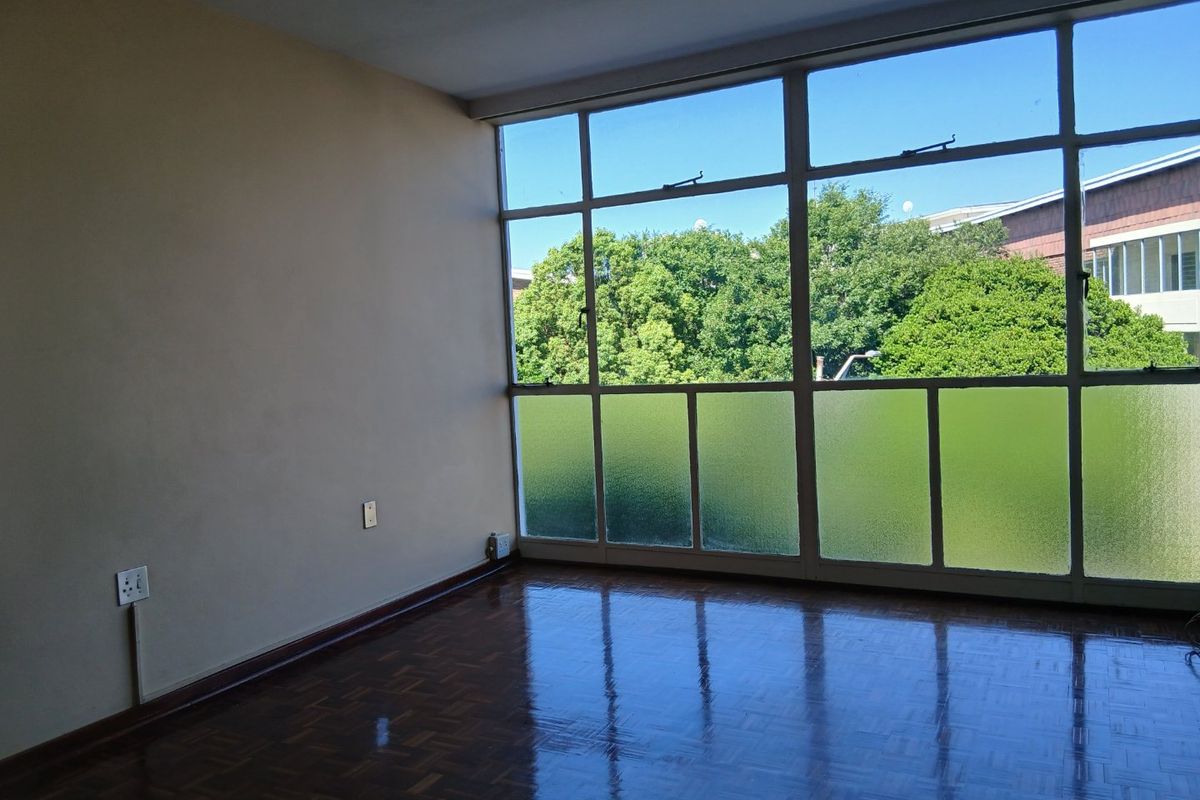 An empty living room featuring polished parquet flooring and large steel-framed windows that provide ample natural light and a pleasant green outlook.