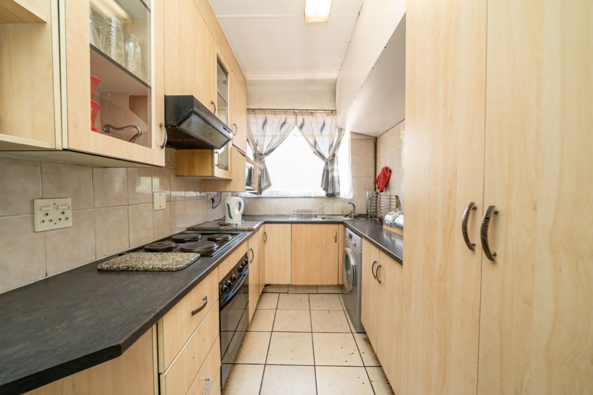 A functional galley-style kitchen featuring light wood-look cabinetry, dark countertops, integrated oven and hob, and tiled flooring with dedicated laundry appliance space.
