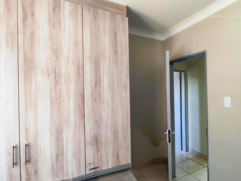 A clean, empty bedroom featuring light wood-grain built-in cupboards, neutral wall tones, and tiled flooring, leading into a connecting hallway.