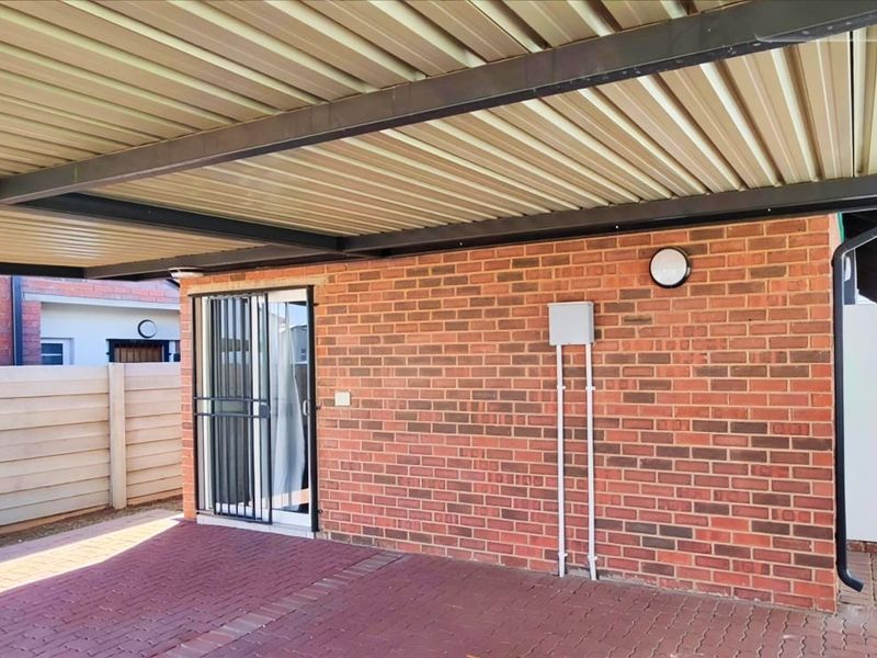 A clean, paved rear exterior area featuring a covered carport, exposed brick wall, and secure access point with a metal security gate.