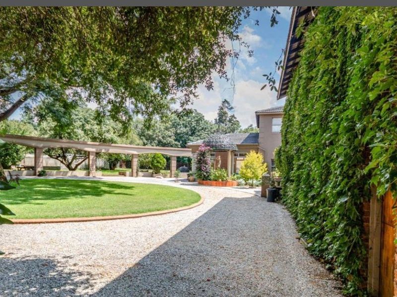 A well-maintained garden featuring a circular lawn, gravel driveway, and a long pergola structure alongside a brick wall covered in climbing ivy.
