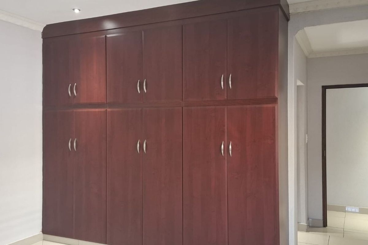 A clean, empty bedroom featuring dark wood built-in cupboards, tiled flooring, and modern recessed downlighting.