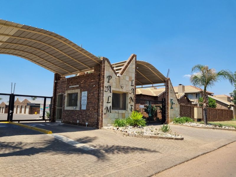 The secure entrance gate and guard house of Palm Lane townhouse complex, featuring brick construction and paved driveway access.