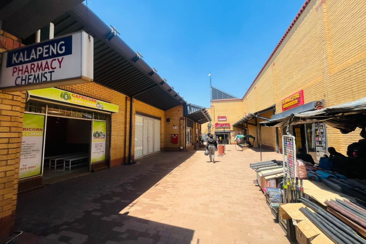 An outdoor pedestrian walkway within a commercial shopping center, featuring brick facades, metal awnings, and various storefront entrances.