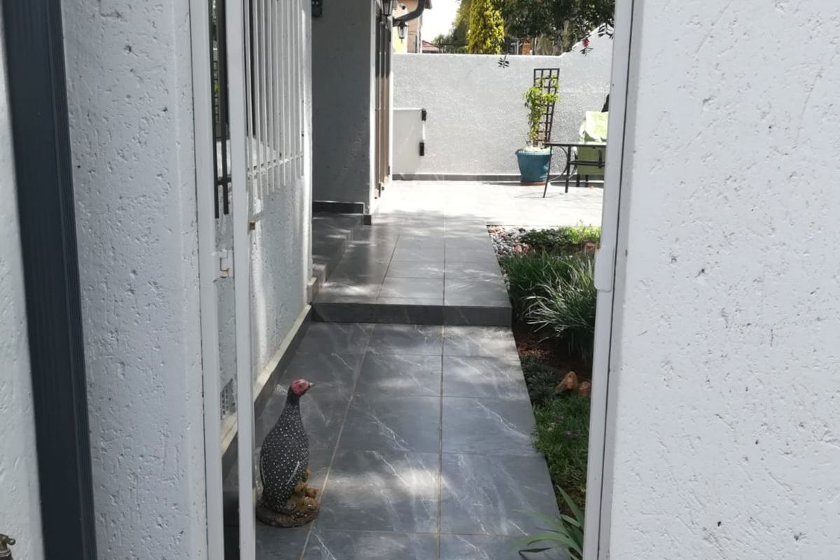 A view through a security gate onto a tiled rear walkway leading to a paved outdoor area and garden space.