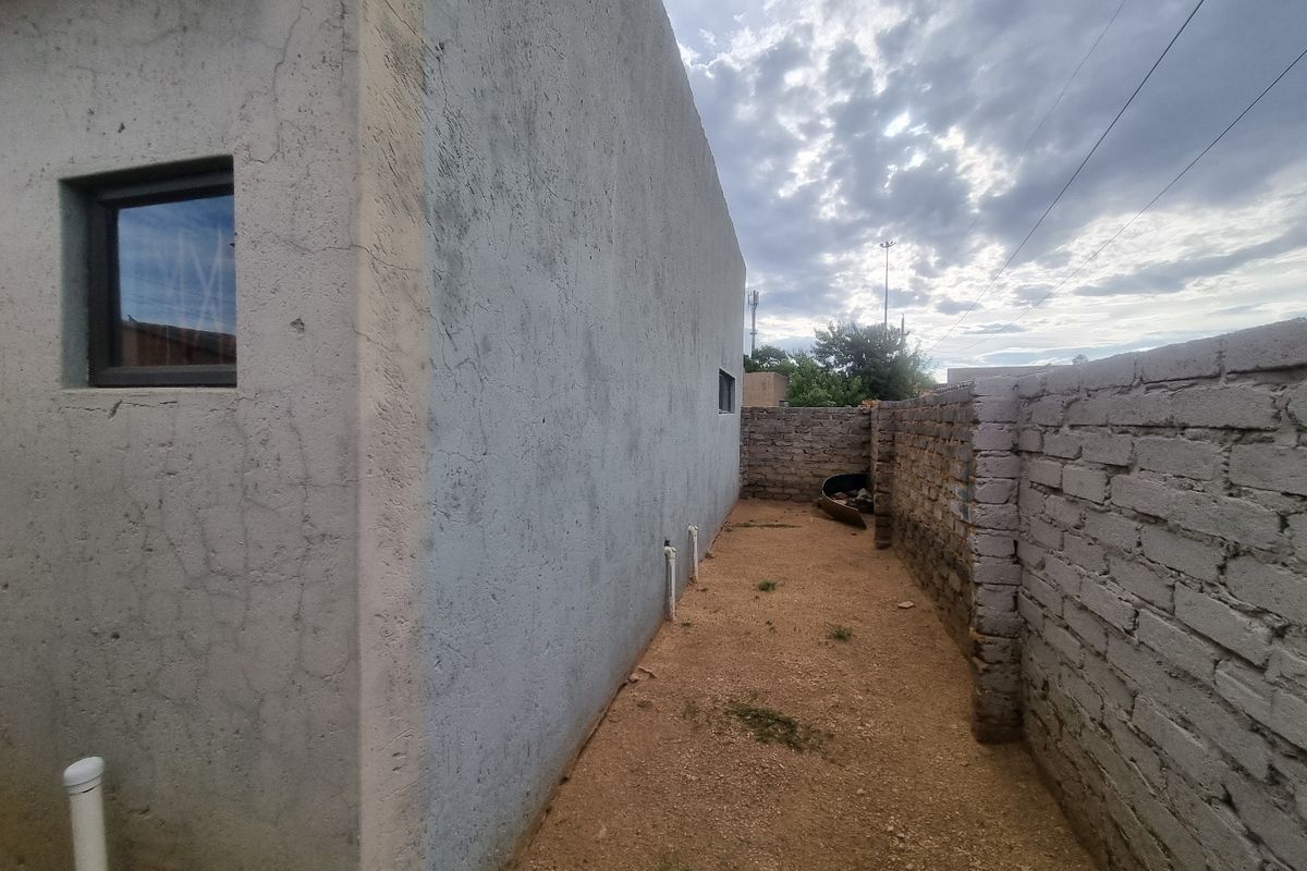 Side exterior view of the commercial property showing a narrow unpaved walkway, exposed brick boundary wall, and exterior plaster finish with visible cracking.