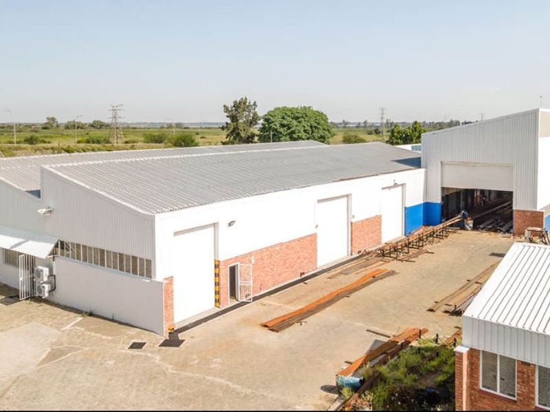 Aerial view of an industrial warehouse facility featuring corrugated metal roofing, brick and plaster exterior, and multiple roller shutter doors with a paved yard.