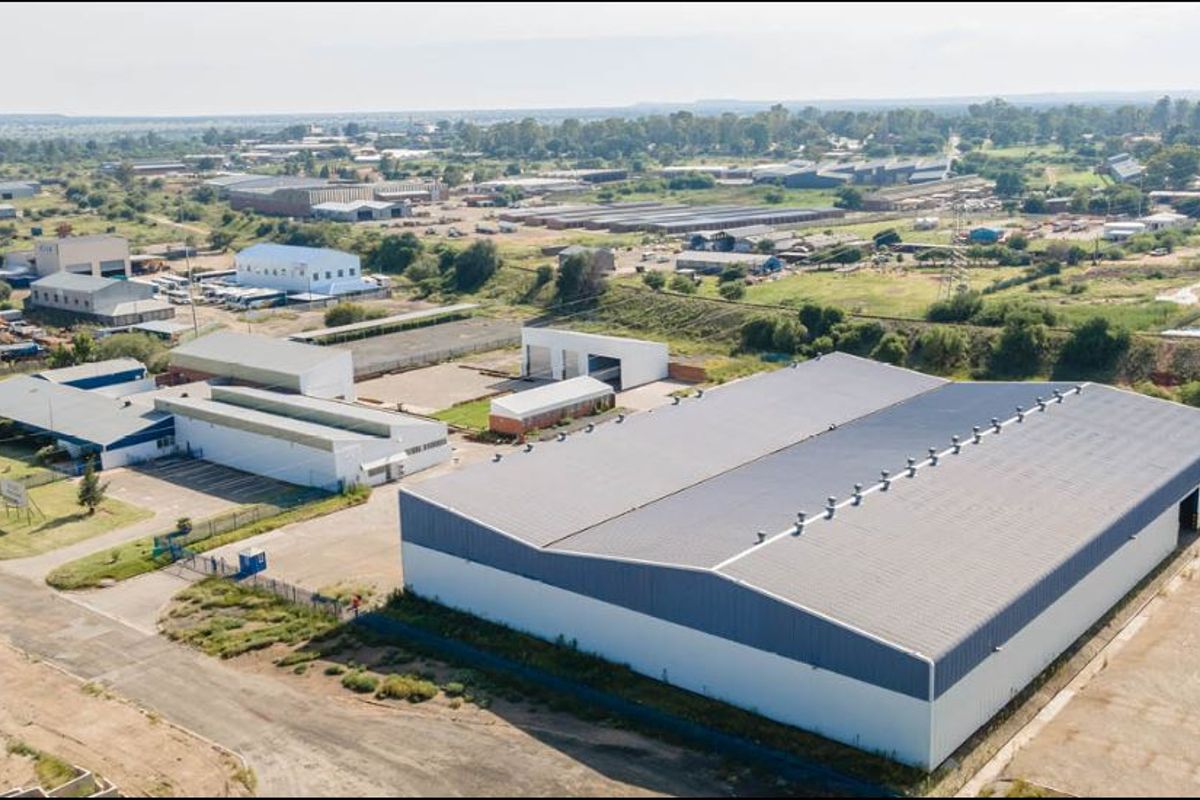 Aerial view of a large industrial property featuring a primary warehouse, multiple auxiliary buildings, and a paved yard within a fenced site.