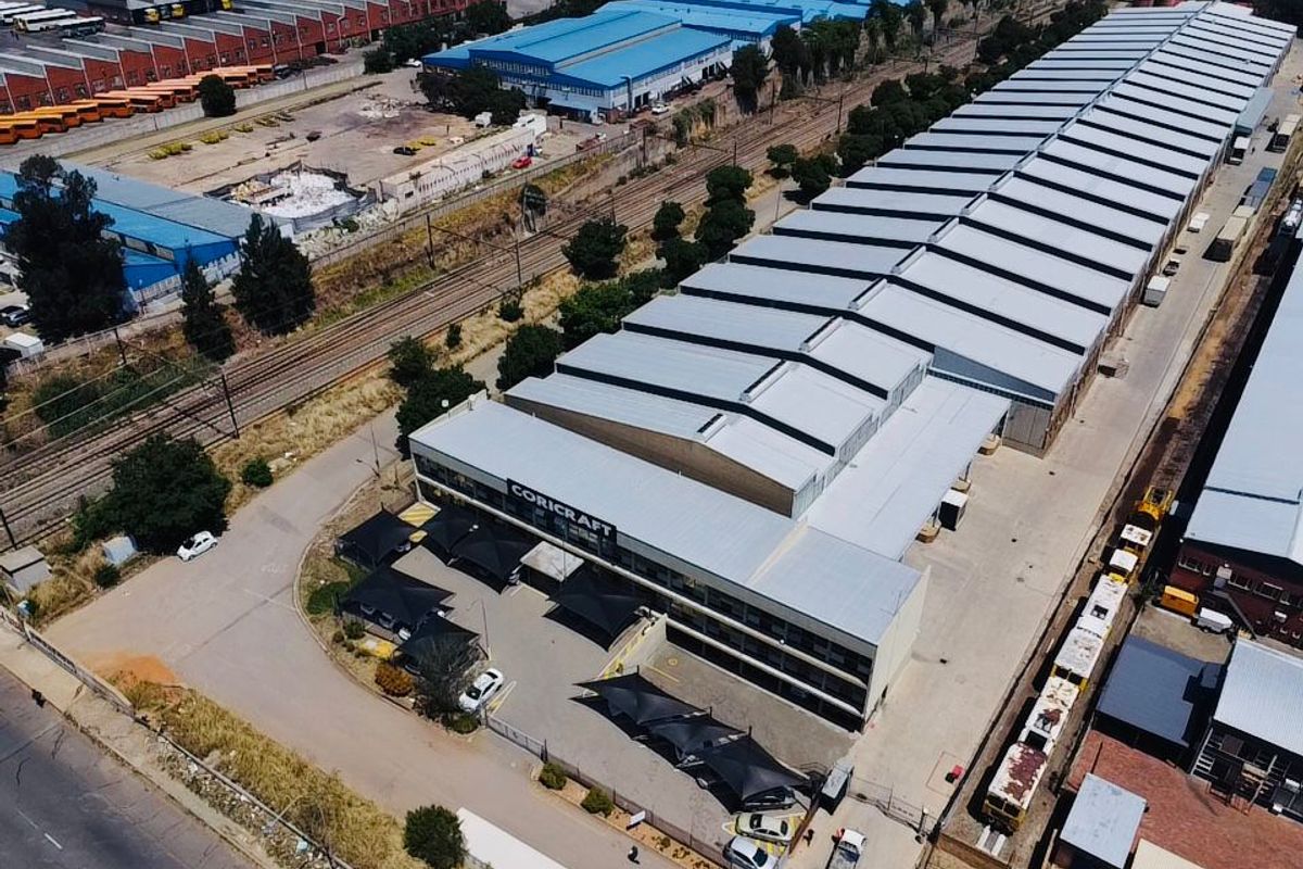 Aerial view of a large industrial warehouse facility featuring a sawtooth roof, extensive paved parking with shade structures, and adjacent railway access.