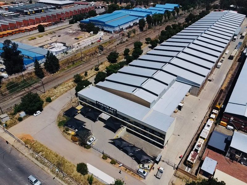 Aerial view of a large industrial warehouse facility featuring a sawtooth roof, extensive paved parking with shade structures, and adjacent railway access.