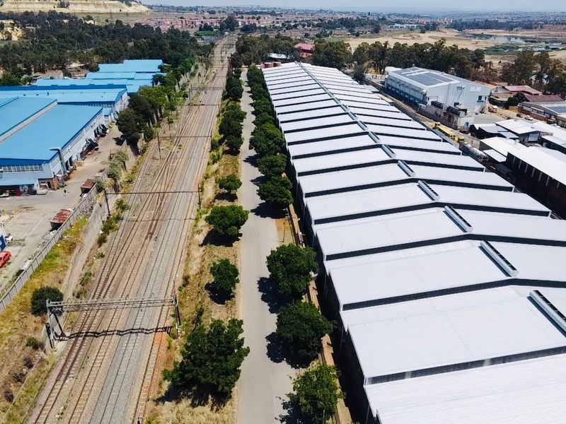 Aerial view of an industrial facility featuring a large warehouse with metal roofing, adjacent to railway tracks and a paved access road.