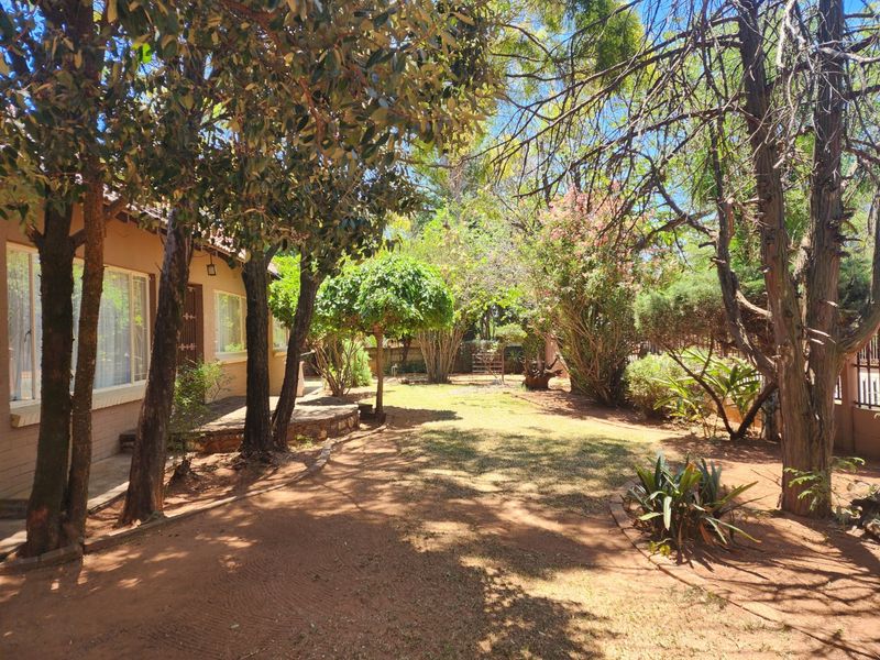 A spacious, established garden featuring mature trees, shaded lawn areas, and a paved patio section adjacent to the house exterior.
