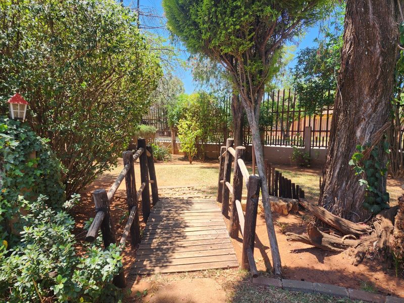 A landscaped garden area featuring a wooden footbridge, mature trees, and secure metal perimeter fencing.