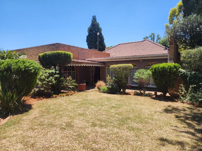 Rear view of a brick house featuring a tiled roof, arched windows, a covered patio area, and a well-established garden.