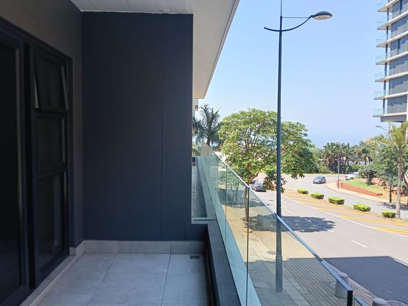 A modern apartment balcony featuring tiled flooring and a sleek glass balustrade, offering an elevated view of the surrounding street and neighborhood.