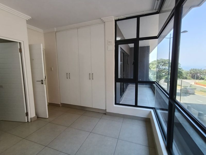 A bright, empty bedroom featuring built-in cupboards, tiled flooring, and large windows providing natural light and access to an adjoining balcony.