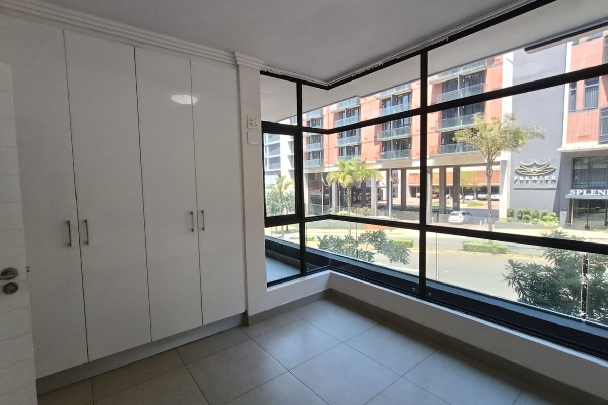 A bright, empty bedroom featuring modern built-in cupboards, large floor-to-ceiling windows with urban views, and neutral tiled flooring.