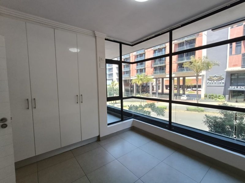 A bright, empty bedroom featuring modern built-in cupboards, large floor-to-ceiling windows with urban views, and neutral tiled flooring.