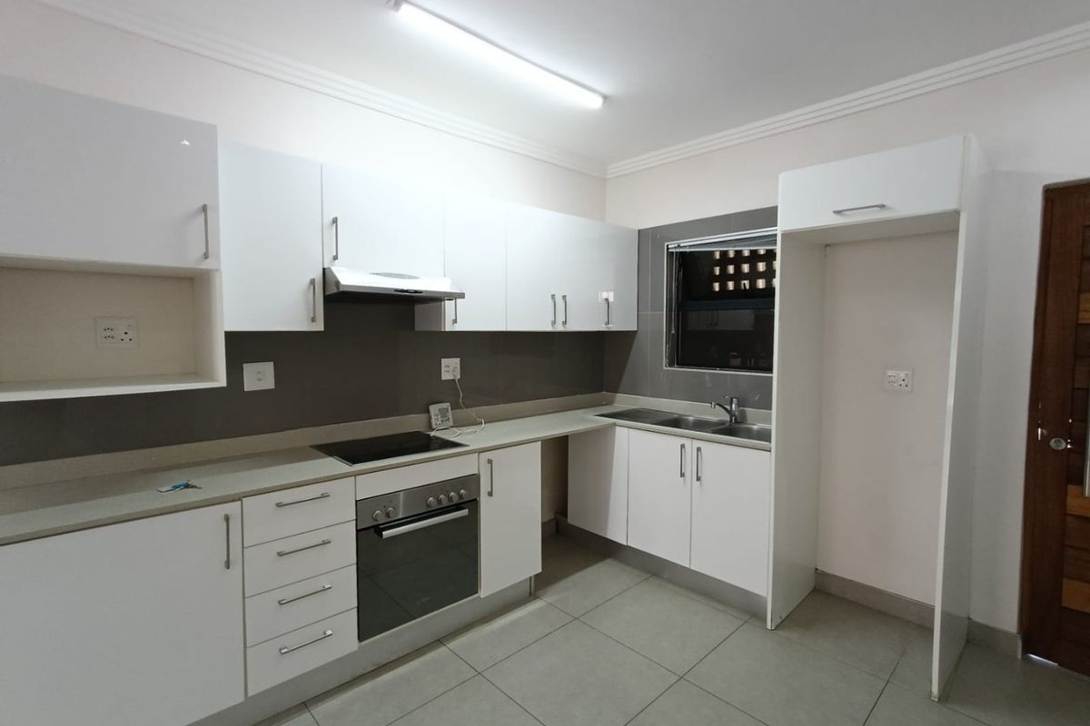 A modern, clean kitchen featuring white cabinetry, stone countertops, integrated oven and hob, and tiled flooring, presented in a vacant, well-maintained condition.