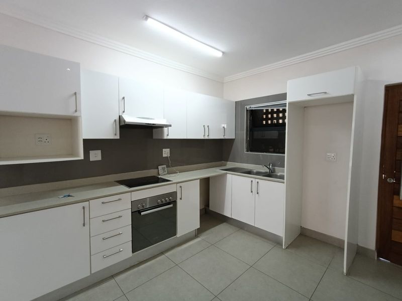 A modern, clean kitchen featuring white cabinetry, stone countertops, integrated oven and hob, and tiled flooring, presented in a vacant, well-maintained condition.